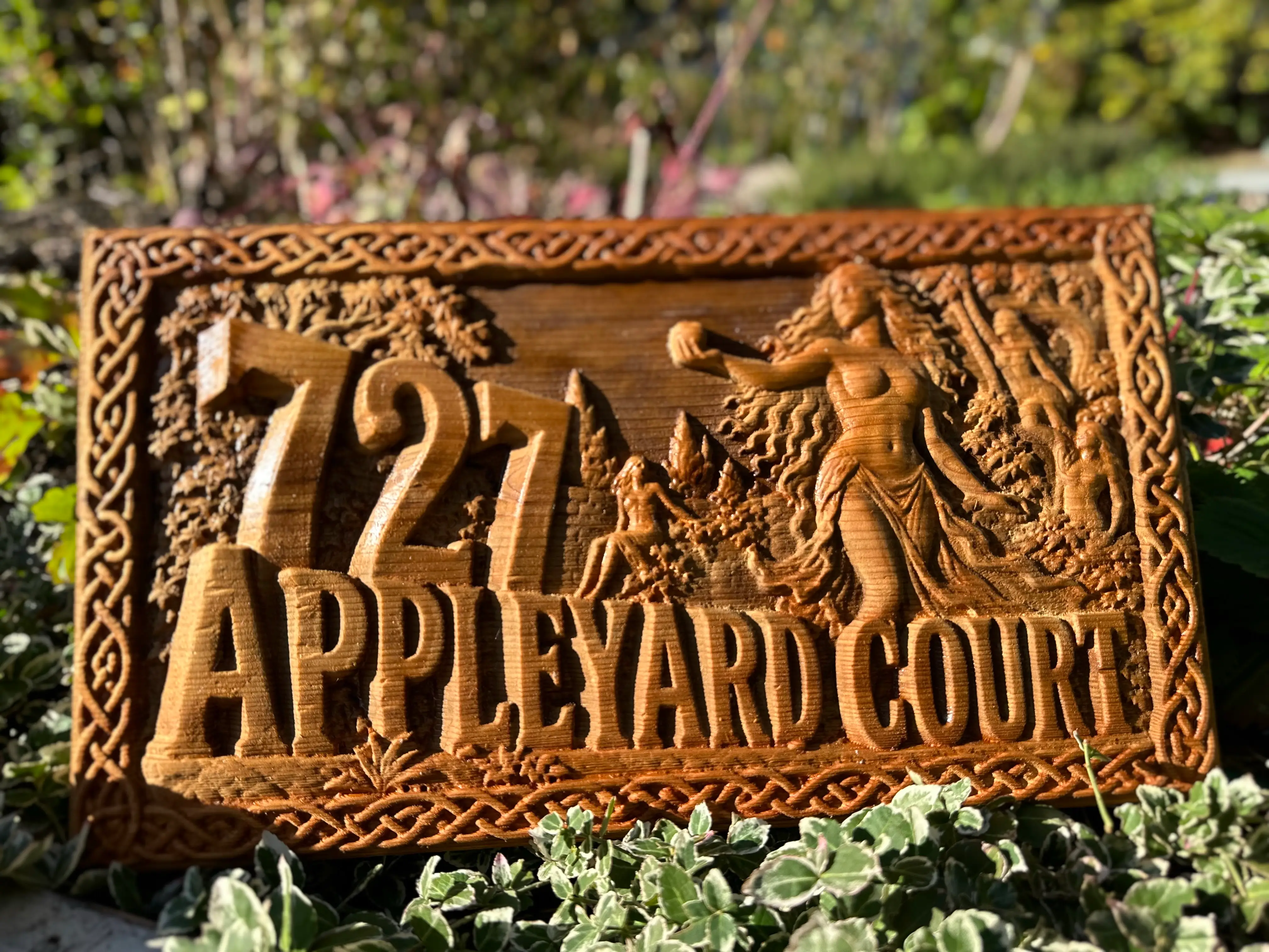 A carved cedar address sign reading 727 Appleyard Court, with a Celtic knot border and a classical garden scene, glowing in low sun among garden plants